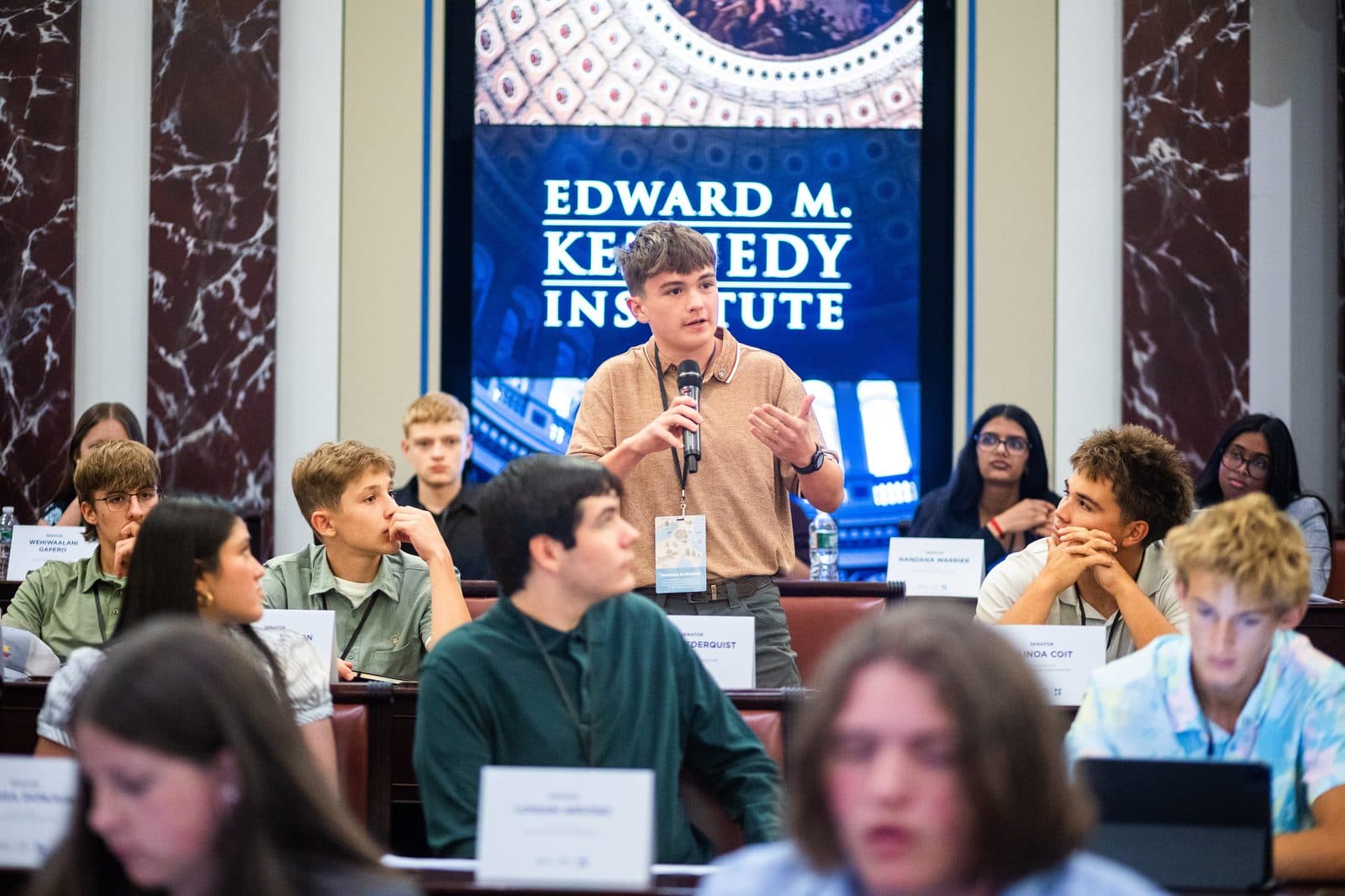 A student speaking into a microphone at the Edward M. Kennedy Institute during a youth debate event.