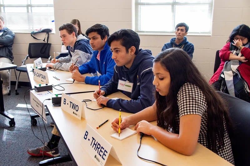 Students at a debate competition, seated at desks labeled with team positions, taking notes.